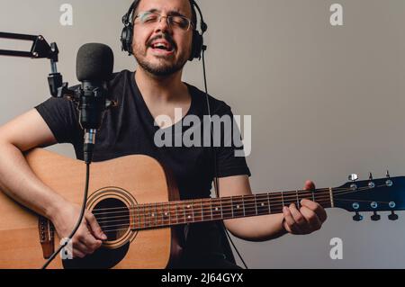 Caucasian young man with beard and glasses, in home studio singing and playing acoustic guitar streaming, recording music for video on social networks Stock Photo