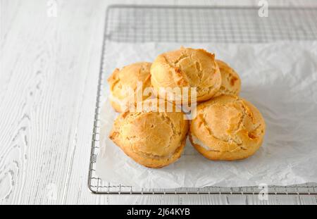 Homemade profiteroles or eclairs on a metal grid. Traditional French dessert. White wooden background. Close-up, copy space. Stock Photo