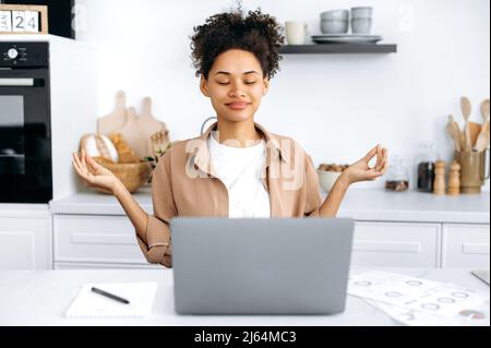 Meditation and relaxation. Happy African American millennial girl, student or freelancer, sits in the kitchen at the table, takes a break from work or study, meditates, relaxes the brain, smiling Stock Photo