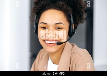 Close-up photo of a confident satisfied young african american woman wearing headphones, call center employee, freelance, assistant, communicating online, looking at camera, smiling friendly Stock Photo