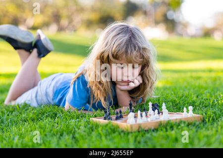 Clever concentrated and thinking child while playing chess, laying on grass in summer park. Chess, success and winning. Stock Photo