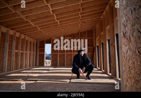 Male engineer building wooden frame house in the Scandinavian style barnhouse. Man developer on construction site, inspecting quality of work on sunny day, holding hammer in hands. Stock Photo