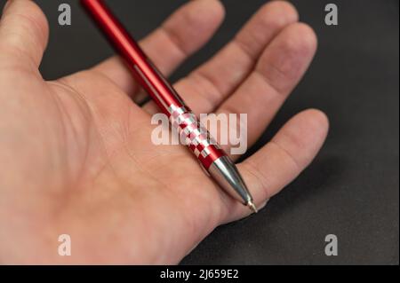 A close-up of a man's hand holding a red pen against a black background. Metal ballpoint pen lying in the open palm of a mature man's hand. Stock Photo
