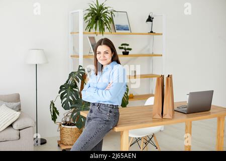 Calm and peaceful young woman takes a break, rests with a cup of hot drink near the table at office. Stock Photo
