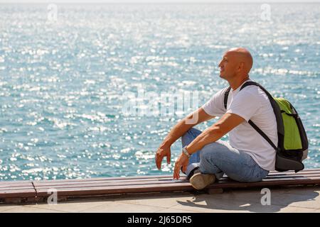Portrait bald man on background sea. Tourist guy sitting on embankment near the pier enjoys a sunny day in casual clothes with a backpack looks around Stock Photo