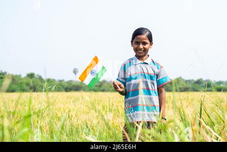 Happy smiling Indian village girl with Indian flag standing in middle of the paddy field by looking at camera - concept of patriotism, repubilc or Stock Photo