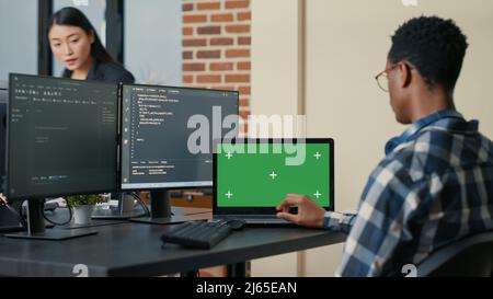 Database designer writing code using laptop with green screen chroma key mockup sitting at desk in busy developers agency. Programer looking at multiple screens with programming language. Stock Photo