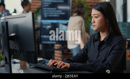 Programer writing algorithm focused on compiling source code sitting at desk in software development office with team working in background. Concentrated system engineer working on parsing data. Stock Photo
