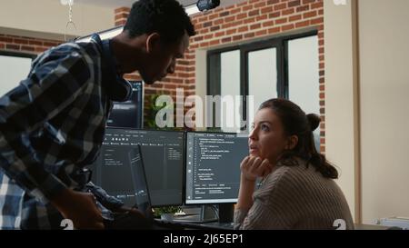 Junior developer sitting down on desk showing laptop with source code to senior dev asking for opinion. Programer writing algorithm interrupted by colleague wanting help with fixing errors. Stock Photo
