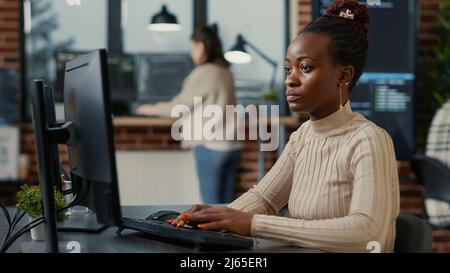 African american software engineer working focused looking at computer screen while typing in it startup office. Database programer writing code with algorithms compiling on screens in background. Stock Photo