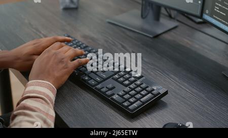 Closeup of african american programer hands typing code on keyboard while looking at computer screens with programming interface. System engineer sitting at desk developing algorithm. Stock Photo