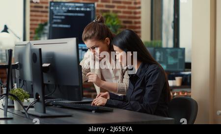 Team of coworkers comparing source codes running on laptop screen and computer monitor in it development office. software developers collaborating on data coding group project while sitting at desk. Stock Photo