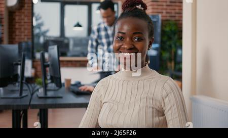 Portrait of african american programer sitting down coding on laptop looking up and smiling at camera. Coder using portable computer next to mixed team in software development agency. Stock Photo
