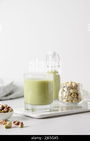 Pistachio milk in glass and pistachios in glass jar on white background. Lactose free. Vegan nutty plant based milk. Vertical. Stock Photo