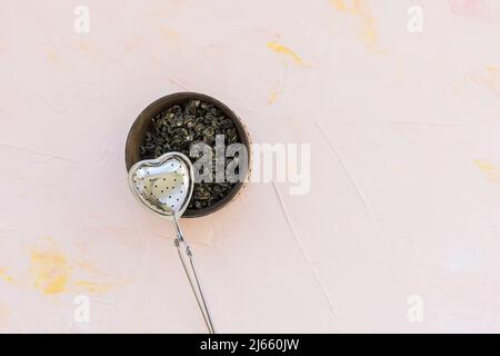 Chinese high quality gunpowder loose leaf tea. Jasmine Green tea leaves rolled in tight spiral. Green snail tea balls in wooden bowl on pink background. Dragon pearl Jasmine Green snail tea balls and tea steeper on background. Top view, copy space Stock Photo