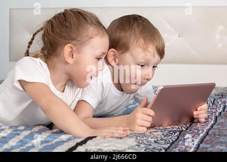 Kids and gadgets. A boy and a girl are lying on the bed and looking at the tablet in surprise Stock Photo