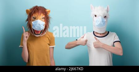 Young woman wearing lion and protection medical mask pointing up with index finger, and in alpaca mask keeps thumb up approval gesture Stock Photo