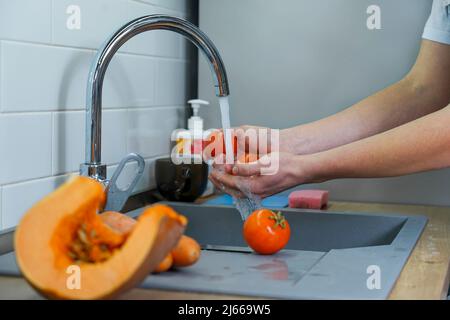 The cook washes fresh vegetables under running water from the tap. He is holding two ripe red tomatoes in his hands Stock Photo