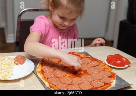 Small girl cooking food. Little child making pizza, sitting at table in kitchen at home, putting pepperoni on dough. Stock Photo