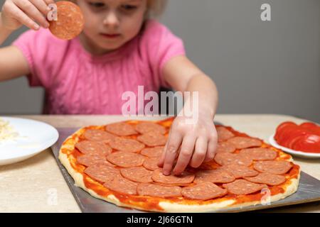Little child making pizza, sitting at table in kitchen at home, putting pepperoni on dough. Small girl cooking food. Stock Photo