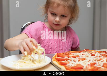 Small child cooking meal at home sitting at the table in the kitchen at home. Little girl making pizza, adding cheese. Stock Photo