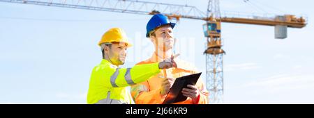 Two Young Male Engineers Standing In Front Of Mechanical Crane Working On The Construction Site Stock Photo