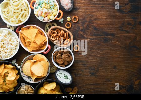 Unhealthy food or snacks. All classic potato snacks with peanuts, popcorn and onion rings and salted pretzels in bowl plates on old wooden background. Stock Photo