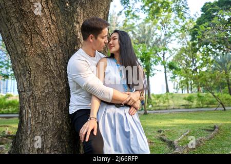 Multiracial couple about to kiss leaning against a tree Stock Photo