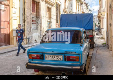 A Cuban man walks in a cobblestone street with weathered buildings on both sides. A blue Russian Lada car is parked in the residential district. Stock Photo