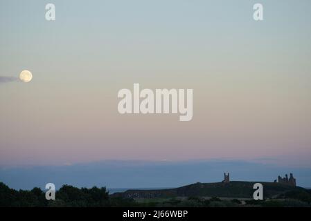 Landscape image of moonrise over Dunstanburgh Castle in early evening autumn light Stock Photo