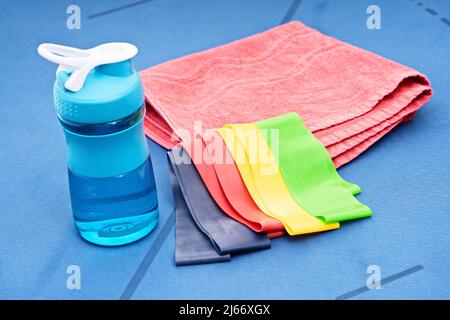 Close up of yoga or fitness items – elastic resistance rubber bands, towel, and water bottle on a blue mat. Sports Concept. Sports accessories.