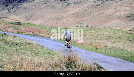 A cyclist starts to rides up Hardknott Pass, one of the steep Lakeland passes which the Fred Whitton Challenge will follow. This is one of the many roads in the Lake District, Cumbria, England, UK, will be closed on May 8th, 2022, when 2,500 cyclists ride the Fred Whitton Challenge route. The Fred Whitton Challenge is a charity cyclosportive event held annually in the English Lake District. It is held in memory of Fred Whitton, racing secretary of the Lakes Road Club, who died of cancer at the age of 50 in 1998. Stock Photo