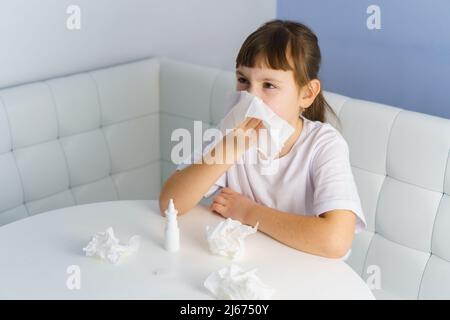 Little girl with runny nose is blowing into paper tissue. Nasal spray and used tissues on table Stock Photo