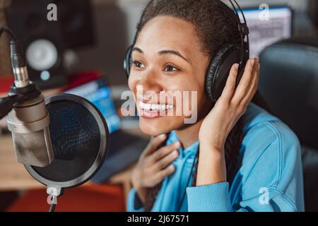 Charming enthusiastic African anchor woman, with headphones talking into microphone in the sound recording studio Stock Photo