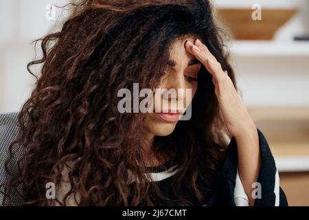 Sad thoughtful curly tanned Latin woman in striped shirt touch forehead look down think about mistakes in work letter essay in home interior. Copy Stock Photo