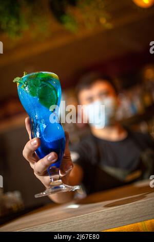 Barman serving blue cocktail on bar counter against dark blurred background. Stock Photo