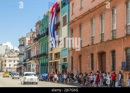 A large group of people waiting for a public omnibus at a bus stop. A large Cuban flag hangs from a government building. Stock Photo