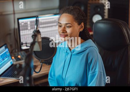 Young multiracial woman blogger, radio presenter, broadcasting into microphone, recording a podcast in broadcast studio Stock Photo