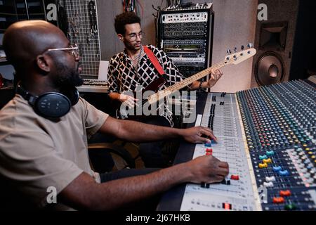 Young Black musician playing guitar and sound engineer adjusting setting on mixer in recording studio Stock Photo