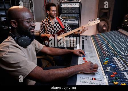 Modern young Black musician playing electric guitar while sound engineer using mixing console in recording studio Stock Photo