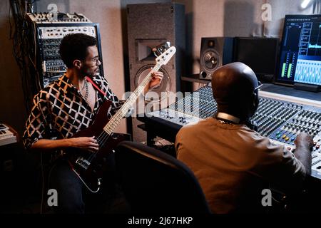 Stylish young Black musician playing electric guitar while music producer working on mixing console in recording studio Stock Photo