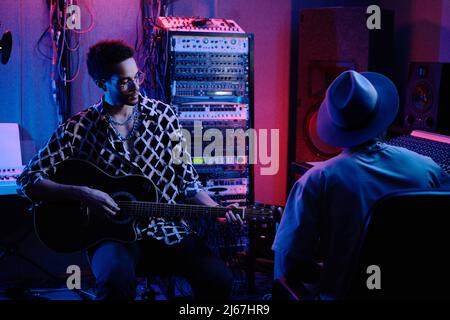Unrecognizable audio engineer and creative young African American guitar player working on album in recording studio, neon lighting Stock Photo