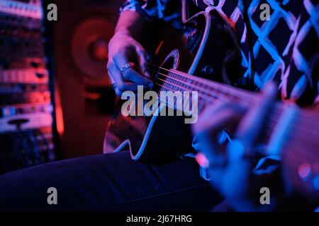 Selective focus shot of unrecognizable young male rock musician playing acoustic guitar in studio, neon lighting Stock Photo