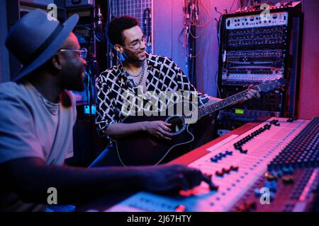 Mature Black music producer and young guitar player enjoying working on album in recording studio, neon lighting Stock Photo