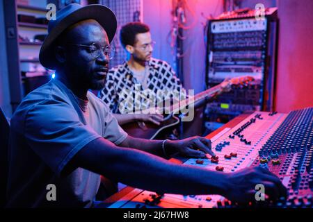 Selective focus shot of stylish mature African American producer adjusting sound on mixer while young musician playing guitar Stock Photo