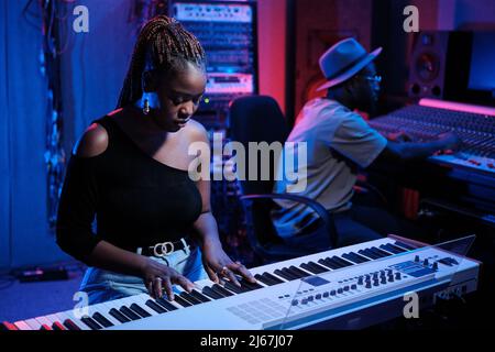 Stylish young African American female musician playing digital piano while audio engineer working on mixer in recording studio Stock Photo