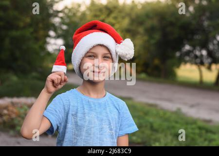 funny smiling boy in Santa's helper hat. in the summer on the street. Against the background of green foliage. Stock Photo