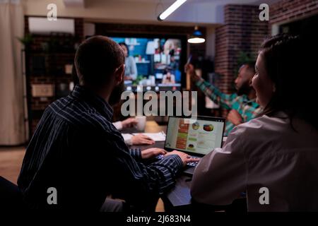 Team in video conference talking with remote colleagues while startup employee is working with sales charts on laptop. Coworkers doing overtime at the office attending internet call meeting. Stock Photo
