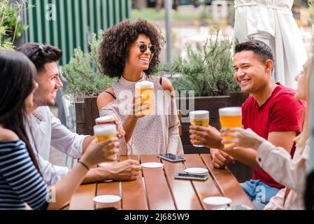 Multi-ethnic group of friends or students sitting down at a wooden table while toasting and drinking beer together outdoors in a pub Stock Photo