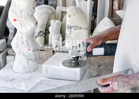 Unrecognizable male artisan with ruler smoothing block of marble with polishing machine during work in sunlit workshop in daytime. Stock Photo